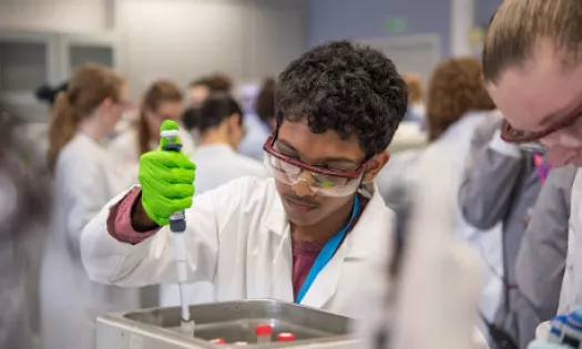 A male student in a lab setting uses a pipette. He is wearing a lab coat, goggles and gloves.